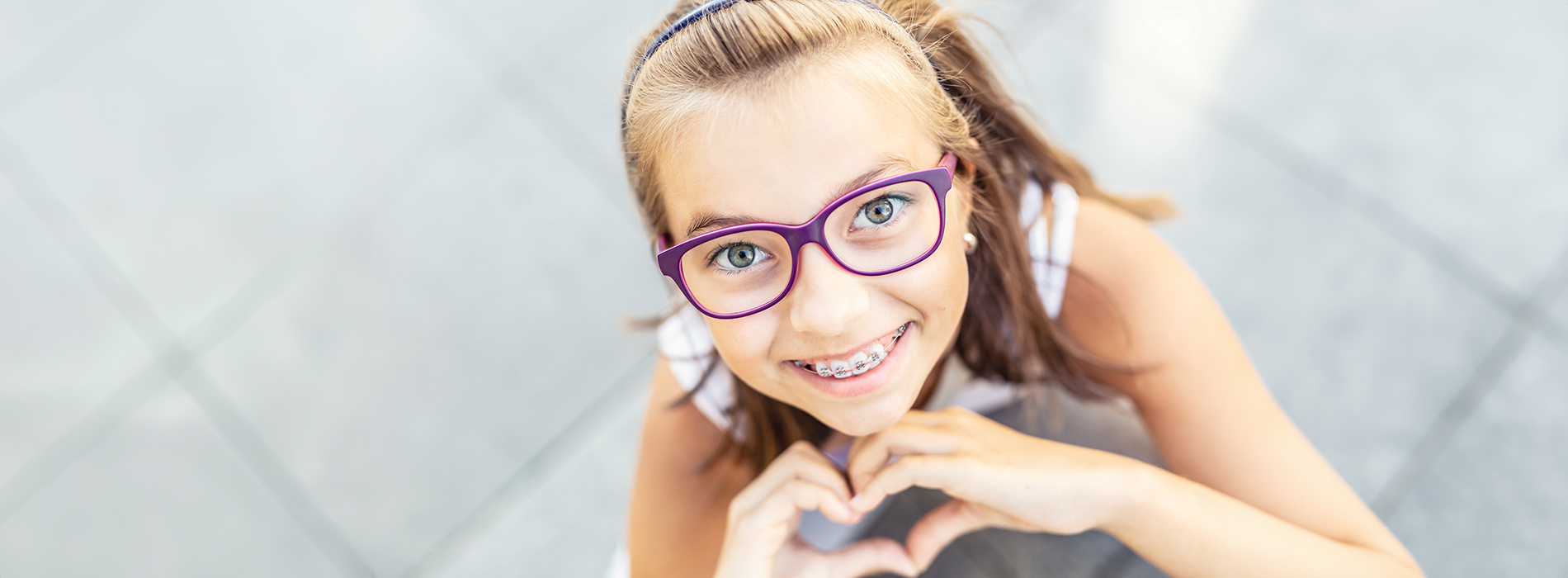 The image features a young girl making a heart shape with her hands, wearing glasses and standing outdoors.