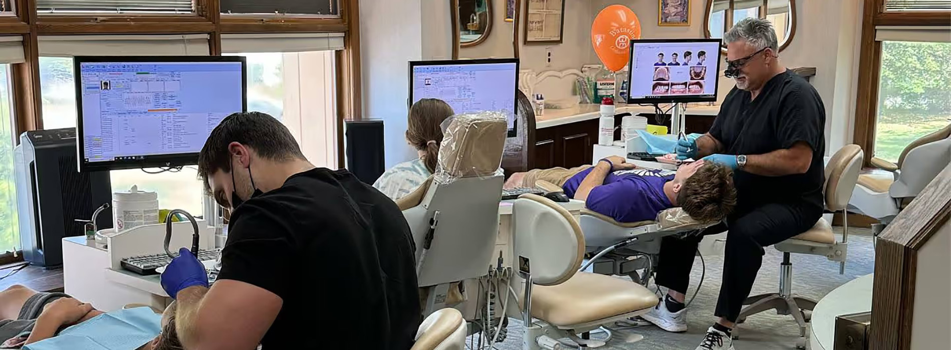 The image shows a dental office with people seated in chairs, receiving dental care  a dentist in a white coat and stethoscope stands at a counter with a computer monitor.