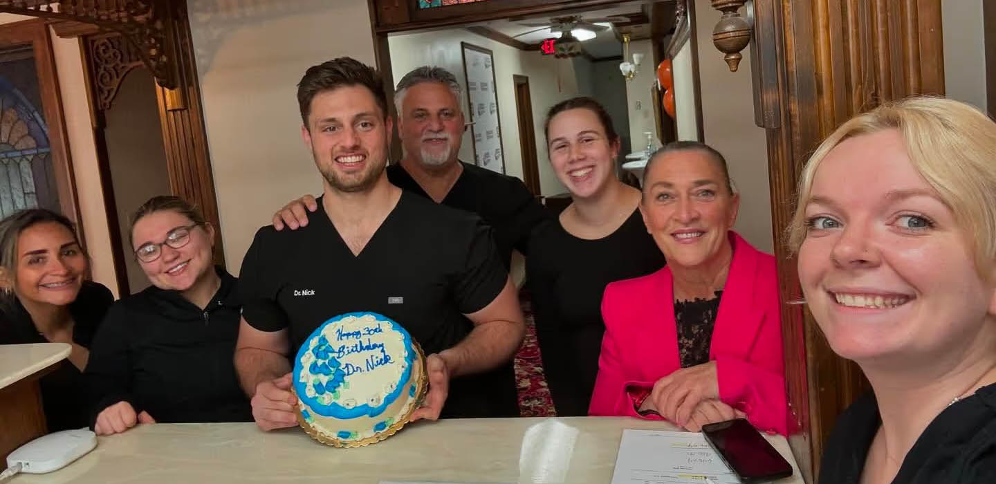 A group of people are gathered around a table with a cake, smiling at the camera.