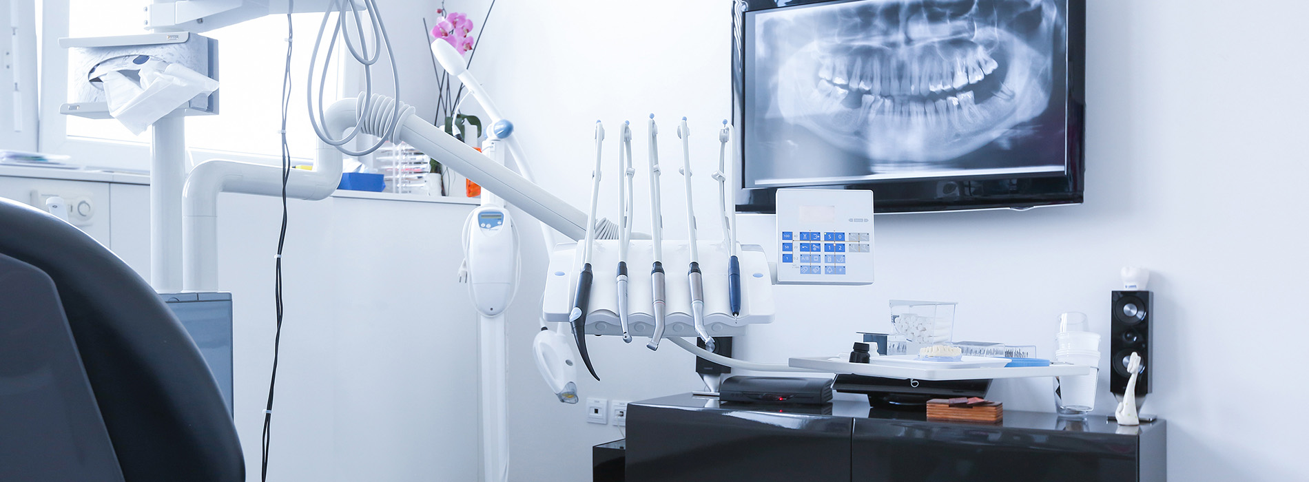 The image shows an interior view of a dental office with dental equipment visible, including a dental chair, a monitor displaying a mouth X-ray, and various dental tools on a countertop.