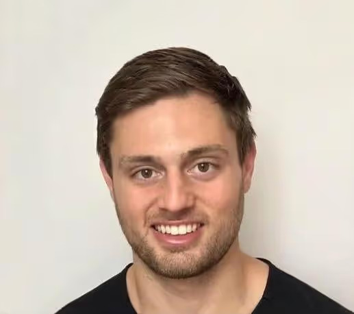 The image shows a smiling man with short hair, wearing a dark top against a white background.