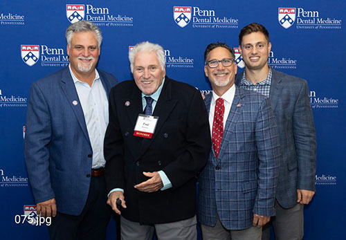 The image shows four men posing together at an event  from left to right  one man wearing a suit with a tie, another wearing glasses and a lanyard, a third man in a suit jacket with a name tag, and a fourth man wearing a plaid blazer and a red tie. They are standing in front of a blue backdrop with text that includes the words  PENN MEDICINE,   WASHINGTON UNIVERSITY SCHOOL OF DENTAL MEDICINE  and  WESTERN NEW YORK.  The men appear to be at a formal event, possibly an awards ceremony or conference.