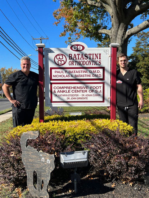 Two men standing in front of a sign with a white background and red lettering.