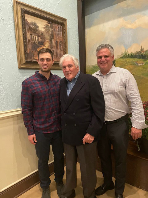 This is a photograph featuring three men posing together at an indoor event. The man on the left is wearing a plaid shirt, the man in the middle is older with gray hair and is wearing glasses, and the man on the right has dark hair and is wearing a suit. They are all smiling and appear to be standing in front of a painting or artwork.