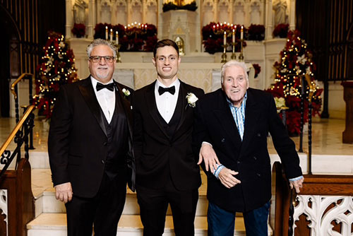 The image shows three men standing together at an altar inside a church during Christmas time, with festive decorations visible in the background.