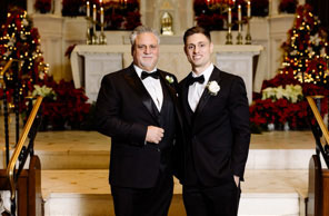 Two men dressed in formal attire, standing together at a church altar during Christmas season, with festive decorations visible in the background.