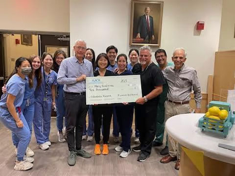 The image shows a group of people gathered around a table with a man holding a check, posing for a photo in an indoor setting, likely a hospital lobby, with a person wearing a surgical mask at the forefront, and a plaque on the wall with a portrait of a man behind them.