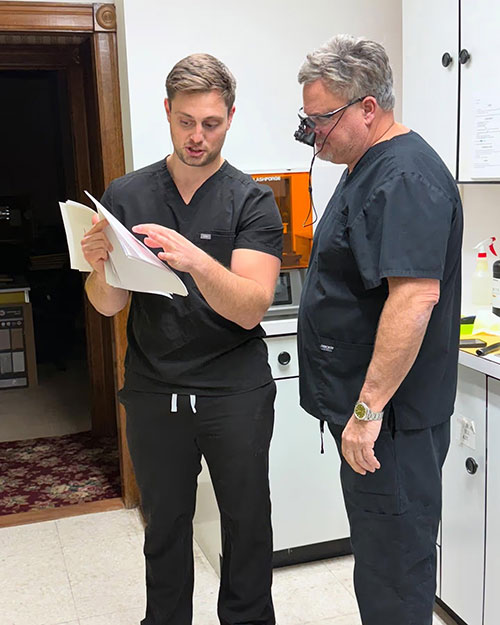 In the image, there are two individuals standing in a room with a countertop and a document being held by one person. The individual on the left appears to be reading the document while the other person is looking at them. Both individuals are wearing scrubs, suggesting they may be healthcare professionals.