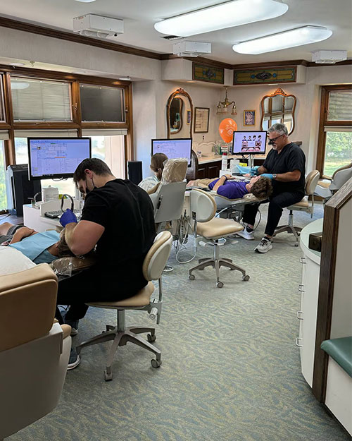 The image shows an interior view of a dental office with multiple chairs equipped for dental procedures, a dentist working at a computer workstation, and various dental equipment visible.