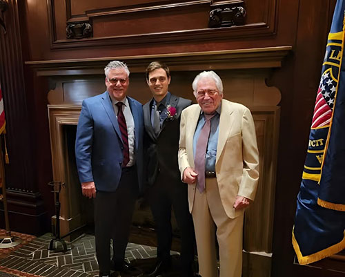 Three men standing together at an event, posing with a flag behind them.
