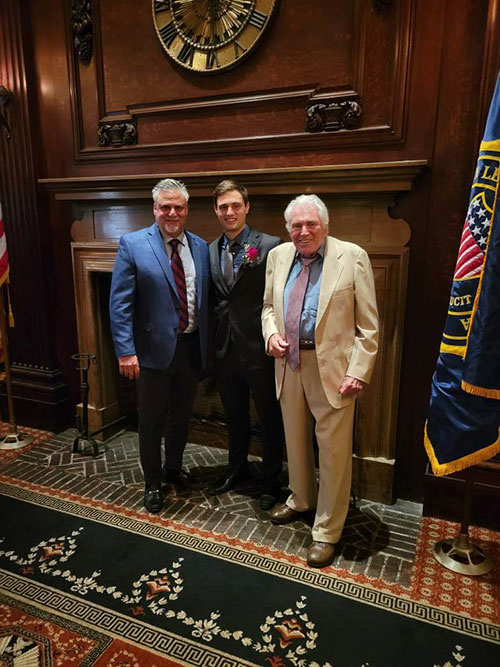 An elderly man, a young man, and another gentleman posing together for a photograph in front of a fireplace with a flag on the wall behind them.