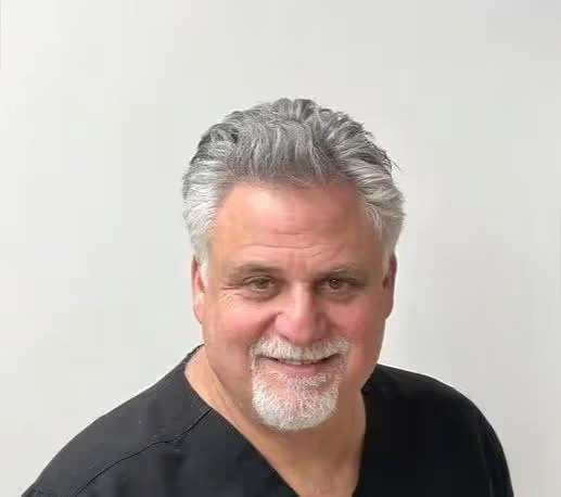 The image shows a man with gray hair and a beard, wearing glasses and a black shirt, standing against a white background. He has a mustache and appears to be smiling slightly.