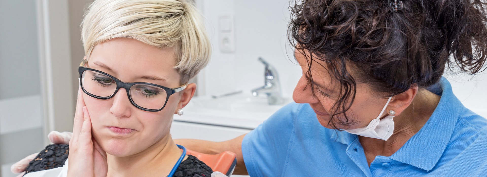 A woman with glasses sits in a dental chair, receiving dental care from a dentist.