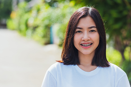 This is a color photograph featuring a person with short hair smiling at the camera. The individual appears to be wearing a white t-shirt and has a tattoo visible on their left arm. They are standing outdoors, possibly in front of a building or garden area, as indicated by the greenery in the background.