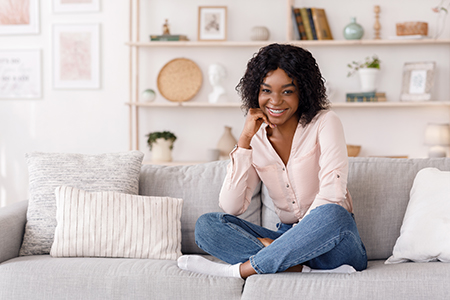 A young woman sitting on a couch with a smile, wearing a pink top and blue jeans, against a backdrop of a home interior.